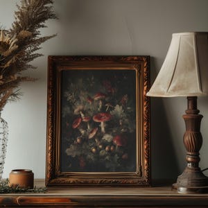 Op de afbeelding: Een ingelijst schilderij met rode paddenstoelen en bladeren, in een donkere, sfeervolle stijl. Het kunstwerk bevindt zich in een sierlijke gouden lijst. De tekst "DARKENDEDBRUSH" staat boven het frame. De schilderij staat op een houten oppervlak.