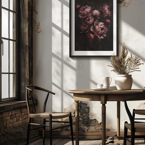 May include: A framed print of a dark floral still life featuring a bouquet of pink peonies against a black background. The peonies are arranged in a vase and appear to be dripping with a dark red liquid. The print is hanging on a white wall in a room with a wooden table and chairs.