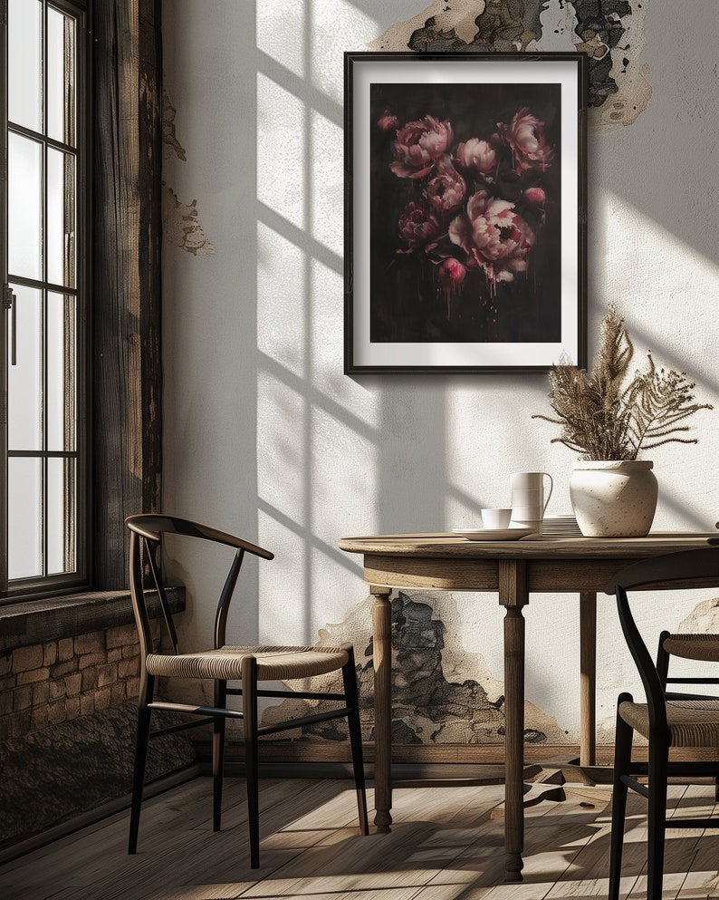 May include: A framed print of a dark floral still life featuring a bouquet of pink peonies against a black background. The peonies are arranged in a vase and appear to be dripping with a dark red liquid. The print is hanging on a white wall in a room with a wooden table and chairs.