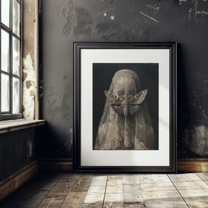May include: A black framed print of a woman wearing a white veil with a large moth covering her face. The background is dark and the image has a vintage feel.