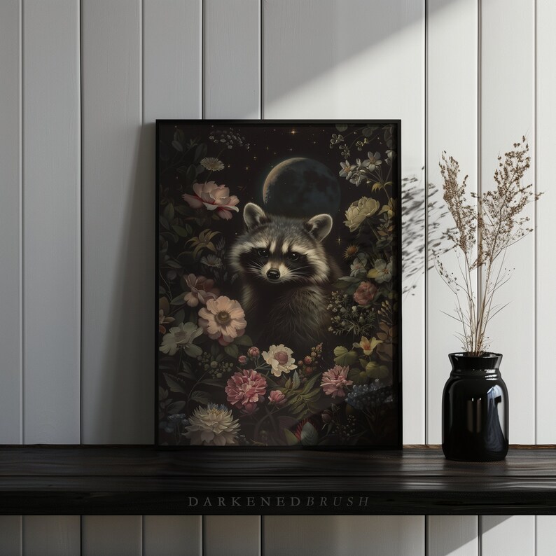 May include: A framed print of a raccoon surrounded by flowers and a crescent moon in a dark, moody style. The raccoon is looking directly at the viewer with a curious expression. The print is set against a white wall with a vase of dried flowers in the foreground.