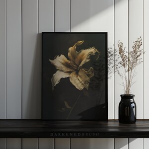 May include: A framed print of a single, dried, light brown lily flower against a dark background. The flower is in focus, with the background blurred. The frame is black.
