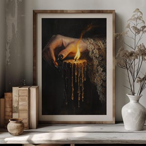 May include: A framed print featuring a hand with long, black nails holding a lit candle. The candle's wax drips down, and the flame glows brightly. The artwork is set against a dark background, with the text "DARKENDEDBRUSH" at the top.