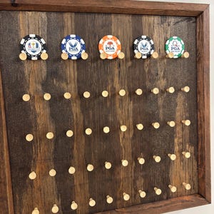 May include: A wooden display board with a dark brown frame and a stained wood background. The board features five golf chip holders with PGA logos and numerous wooden pegs for additional chip storage. The chips are black, blue, orange, and green.
