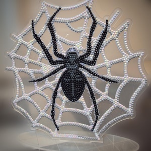 May include: A decorative spider and web ornament crafted from iridescent white and black faceted beads on a clear acrylic stand. The spider is positioned in the center of the web, featuring a black body and eight long, slender black legs.