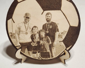 Puede incluir: Un balón de fútbol de madera con una imagen grabada con láser de cuatro personas, incluyendo dos adultos y dos niños, todos con medallas. El balón de fútbol está en un soporte de madera.