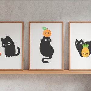 May include: Three framed illustrations of black cats with different accessories. The first cat is holding a slice of watermelon. The second cat is wearing an orange on its head. The third cat is holding a pineapple.