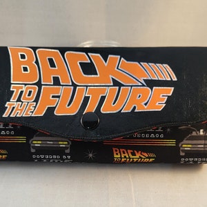 May include: Black wallet with a snap closure. The front features the words "Back to the Future" in orange and white. The back features a repeating pattern of the DeLorean time machine and the words "Back to the Future" in orange and white.