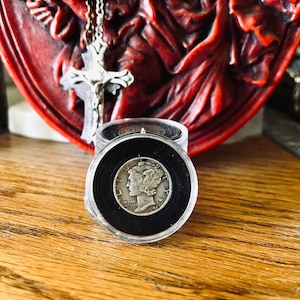 Könnte beinhalten: Eine silberne Münze mit einem Profil eines Frauenkopfes auf der einen Seite, in einem schwarzen Münzhalter. Die Münze befindet sich in einem durchsichtigen Plastiketui mit einem schwarzen Deckel.