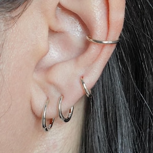 Puede incluir: Una oreja con varios pendientes de aro de plata. Los pendientes son de diferentes tamaños y se llevan en el lóbulo y el helix.