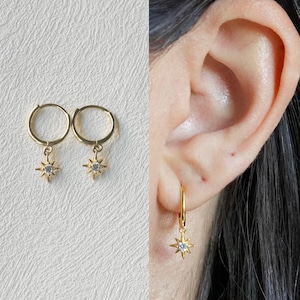 Puede incluir: Un par de pendientes de aro dorados con un colgante de estrella que presenta un cristal transparente en el centro. Los pendientes se muestran en la oreja de una modelo.