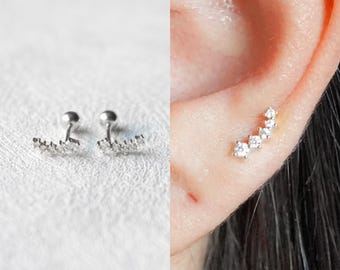 May include: Silver stud earrings with a curved design, each featuring a line of small, clear gemstones. The earrings are shown on a white surface and also worn on an ear.