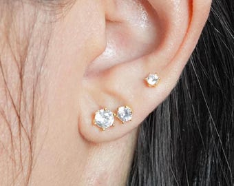 Può includere: Tre piccoli orecchini rotondi di zircone cubico trasparente incastonati in oro. Gli orecchini sono indossati sul lobo dell'orecchio e sulla cartilagine superiore.