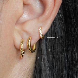 May include: Three gold hoop earrings with different designs. The top earring has a row of small clear stones. The middle earring is a smooth hoop. The bottom earring has a textured design.