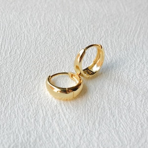 Puede incluir: Un par de pendientes de aro dorados. Los pendientes son peque&ntilde;os, con un dise&ntilde;o redondeado y pulido. Los aros se muestran sobre una superficie blanca texturizada.