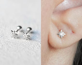 May include: A pair of silver star-shaped earrings with a small clear stone in the centre. The earrings are shown on a white background and on a person's ear.