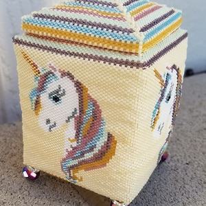 May include: A small, square, beaded box with a lid. The box is decorated with a unicorn design and has a striped lid. The box has four feet made of iridescent beads.