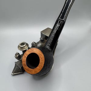 May include: A black tobacco pipe with a wooden bowl and a black stem. The pipe has a textured finish and a silver band. The pipe is resting on a small, decorative metal object.