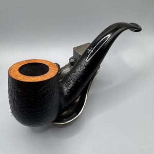 Peut inclure: Une pipe à tabac noire avec un tuyau incurvé et un foyer texturé. Le foyer a un bord brun clair. La pipe repose sur un support de couleur argentée. Une petite pièce métallique est fixée à la pipe.