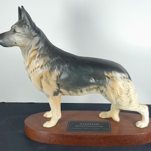 May include: A ceramic Alsatian German Shepherd Dog figurine. The dog is depicted in a standing pose, with a black and tan coat, and is mounted on a wooden oval base. A small plaque with text is affixed to the base.