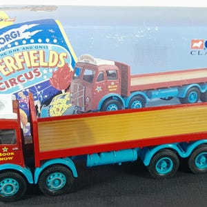 Puede incluir: Un camión de juguete Corgi Chipperfields Circus rojo y amarillo con ruedas azules. El camión tiene las palabras "Book Now" en el lateral. La caja detrás del camión tiene una imagen del camión y las palabras "Corgi Classics".