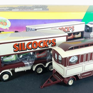 May include: A selection of vintage toy trucks and trailers. The main truck is maroon and white, branded "SILCOCK'S" with "Wigan" on the side. Other trailers include "JACK'S" and a white and maroon trailer. The box reads "The Showman's Range".