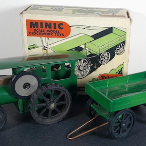 Könnte beinhalten: Ein grüner und schwarzer Spielzeug-Dampftraktor im Vintage-Stil mit passendem Anhänger und Originalverpackung. Auf der Box steht "MINIC SCALE MODEL CLOCKWORK TOYS". Der Traktor hat große schwarze Räder und einen Schornstein.