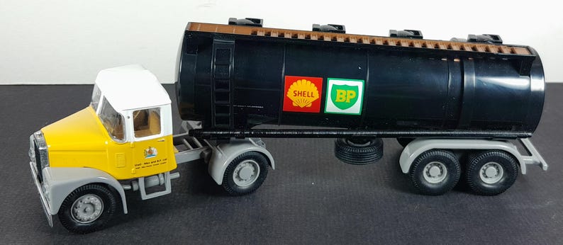Op de afbeelding: Een gele en witte speelgoed vrachtwagen met een zwarte tankwagen. De tankwagen heeft Shell- en BP-logo's. De vrachtwagen heeft grijze spatborden en wielen. De cabine heeft een wit dak.