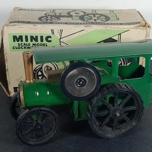 Könnte beinhalten: Ein grüner und schwarzer Spielzeug-Dampftraktor mit dem Text "MINIC SCALE MODEL CLOCKWORK". Das Spielzeug hat große schwarze Räder und einen goldfarbenen Schornstein. Der Traktor steht vor einer Pappschachtel mit einer ähnlichen Traktorillustration.