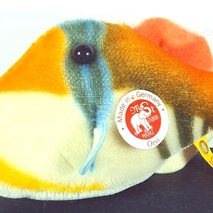 May include: A plush toy fish with a vibrant color scheme. The toy features a mix of orange, yellow, blue, and green hues. It has a round tag that says "Made in Germany" and a yellow tag attached.