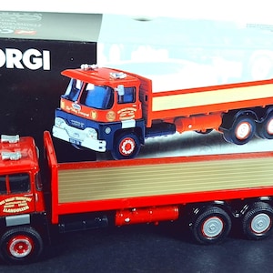 May include: A red and orange Corgi flatbed truck model with a blue cab. The model has a detailed design, including the cab, flatbed, and wheels. The box behind the truck has the Corgi logo and text. The truck model is a collectible item.