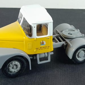 Op de afbeelding: Een gele en witte speelgoedvrachtwagen met grijze accenten. De cabine is geel met een wit dak en grijze spatborden. De vrachtwagen heeft zwarte banden en een trekhaak. De zijkant van de vrachtwagen heeft een logo en tekst.