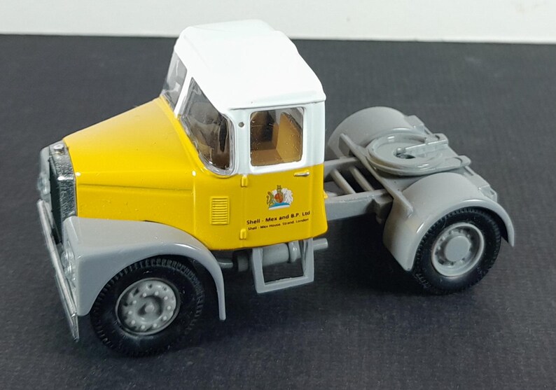 Op de afbeelding: Een gele en witte speelgoedvrachtwagen met grijze accenten. De cabine is geel met een wit dak en grijze spatborden. De vrachtwagen heeft zwarte banden en een trekhaak. De zijkant van de vrachtwagen heeft een logo en tekst.