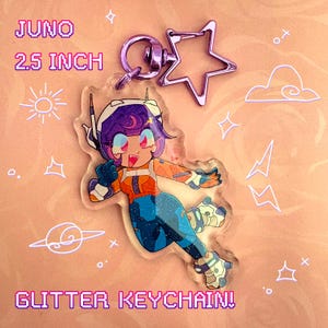 Op de afbeelding: Een glitter sleutelhanger met een stripfiguur genaamd Juno, 6,35 cm hoog. Het personage heeft paars haar, een oranje en blauwe outfit en is bevestigd aan een stervormige sluiting. De sleutelhanger heeft de tekst "Glitter Keychain!"