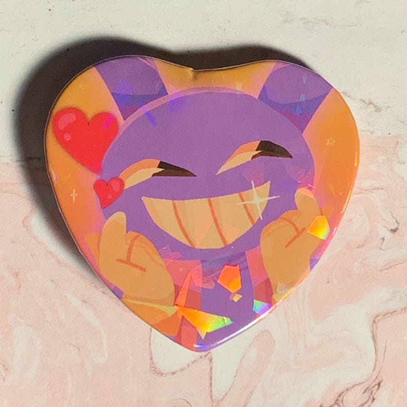 May include: A heart-shaped button featuring a purple character with a wide smile and sparkling eyes. The character is surrounded by a peach background with a red heart and sparkles.
