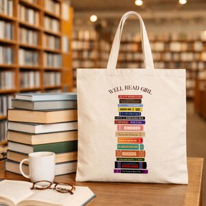 May include: Cream-colored tote bag with the text "WELL READ GIRL" and a stack of book spines. The book spines are in various colors and feature book titles. A stack of books, a mug, and glasses are on a wooden surface.