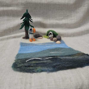 Puede incluir: Escena de paisaje afieltrado con un frailecillo, un árbol y una pequeña cueva. La escena está sobre una base de fieltro azul y verde, con un fondo neutro. El frailecillo tiene patas y pico naranjas. El árbol es verde.