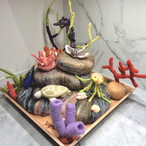 May include: A felted ocean diorama featuring a variety of sea creatures and coral. The scene includes a seahorse, stingray, clownfish, turtle, and octopus, with colorful coral and seaweed accents. The diorama is displayed on a wooden tray.