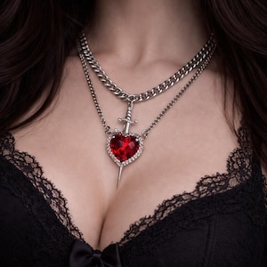 May include: A silver necklace featuring two chains. The upper chain is a chunky curb chain, and the lower chain supports a pendant. The pendant is a red heart-shaped gem, encircled by clear crystals, with a dagger through it.