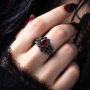 Puede incluir: Un juego de anillos negros con un diseño floral y una gema roja redonda y grande. El anillo está en un dedo con esmalte de uñas rojo oscuro. El anillo tiene una banda a juego con un diseño floral similar.