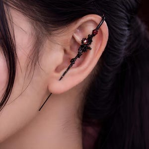 Puede incluir: Puño de oreja negro con un diseño de filigrana adornado. El puño presenta detalles de piedras preciosas rojas y una barra larga y recta. La joya se lleva en la oreja.