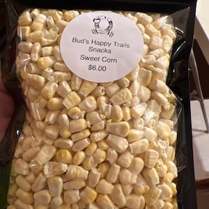 May include: A clear plastic bag filled with sweet corn kernels. A white circular label reads "Bud's Happy Trails Snacks Sweet Corn". The kernels are a mix of yellow and white.