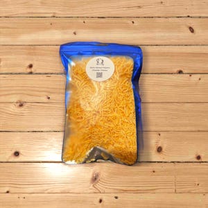 May include: A blue resealable bag filled with shredded cheddar cheese. The bag has a white label with the text "Ruth's Kitchen Helpers Cheddar Cheese" and a QR code.