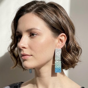 May include: A pair of drop earrings featuring two stacked rectangular metal plates with a textured, hammered surface. The top rectangle is silver, while the bottom rectangle transitions from a pale green to a vibrant blue gradient finish.