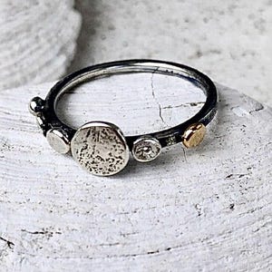 May include: A thin silver ring featuring a series of small, circular accents along the band. The design includes a larger, textured circular centerpiece, a smaller smooth silver disc, and a single polished gold-toned circular accent piece.
