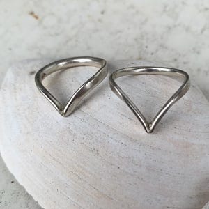 Op de afbeelding: Twee zilverkleurige ringen met een V-vormig chevronontwerp. Elke ring heeft een gladde, gepolijste metalen band die in het midden toeloopt in een punt, wat zorgt voor een opvallend hoekig silhouet. Het metaal heeft een glanzende afwerking.