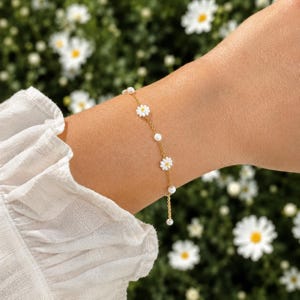 May include: A gold chain bracelet featuring alternating white daisy flower charms with yellow centers and small white spherical beads. The delicate jewelry piece is worn on a wrist, showcasing a repeating pattern of floral and bead elements.