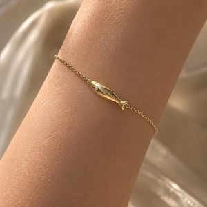 May include: A delicate gold chain bracelet featuring a small, smooth, polished gold fish charm as the central element. The fish charm is accented with a single, tiny, clear round gemstone set into the body, creating a subtle, refined look.