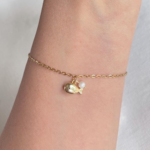 May include: A delicate gold-tone chain bracelet featuring a small, polished gold-tone whale charm and a single, tiny white spherical bead. The bracelet is designed with a simple link chain that secures around the wrist with a metal clasp.