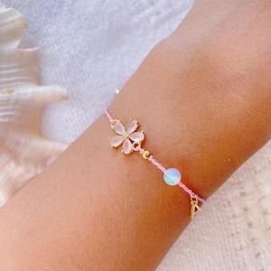 May include: A delicate pink cord bracelet featuring a gold-toned flower charm with translucent petals, a small gold-toned bead, a round iridescent bead, and a gold-toned letter A charm dangling from the string. The bracelet is tied around a wrist.